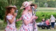 Trois femmes en train de chanter avec robes et chapeaux fleuris et des personnes en train de manger en arrière-plan avec des vignes